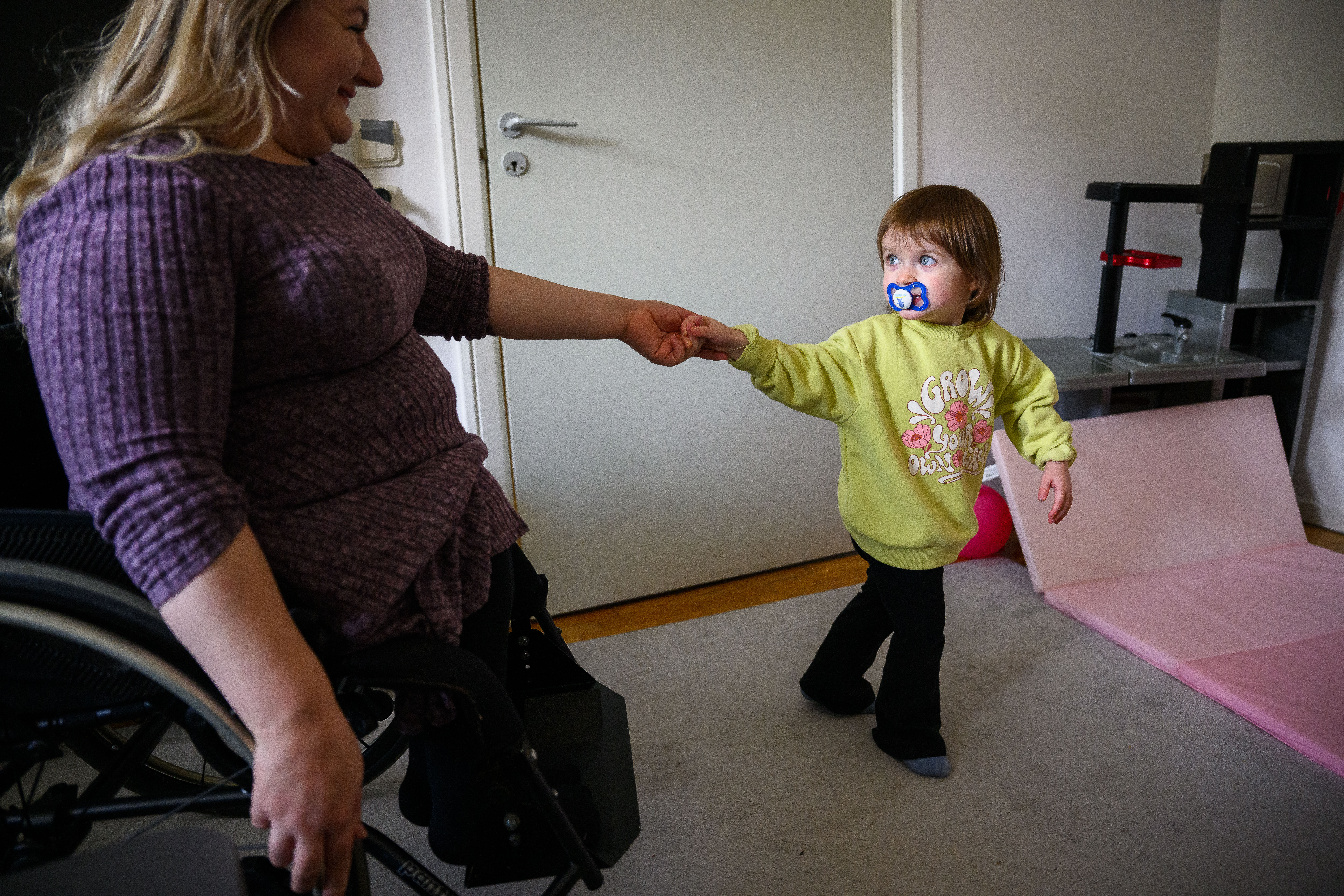 En mamma som sitter i rullstol håller ett litet barn i handen och ler mot barnet. Barnet har en napp i munnen och tittar tillbaka på mamman 