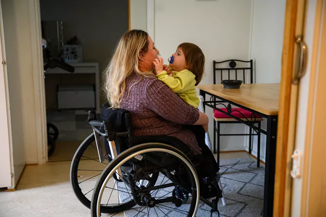 En mamma som sitter i rullstol och har ett barn i knäet. Barnet och mamman kramas och tittar på varandra. 