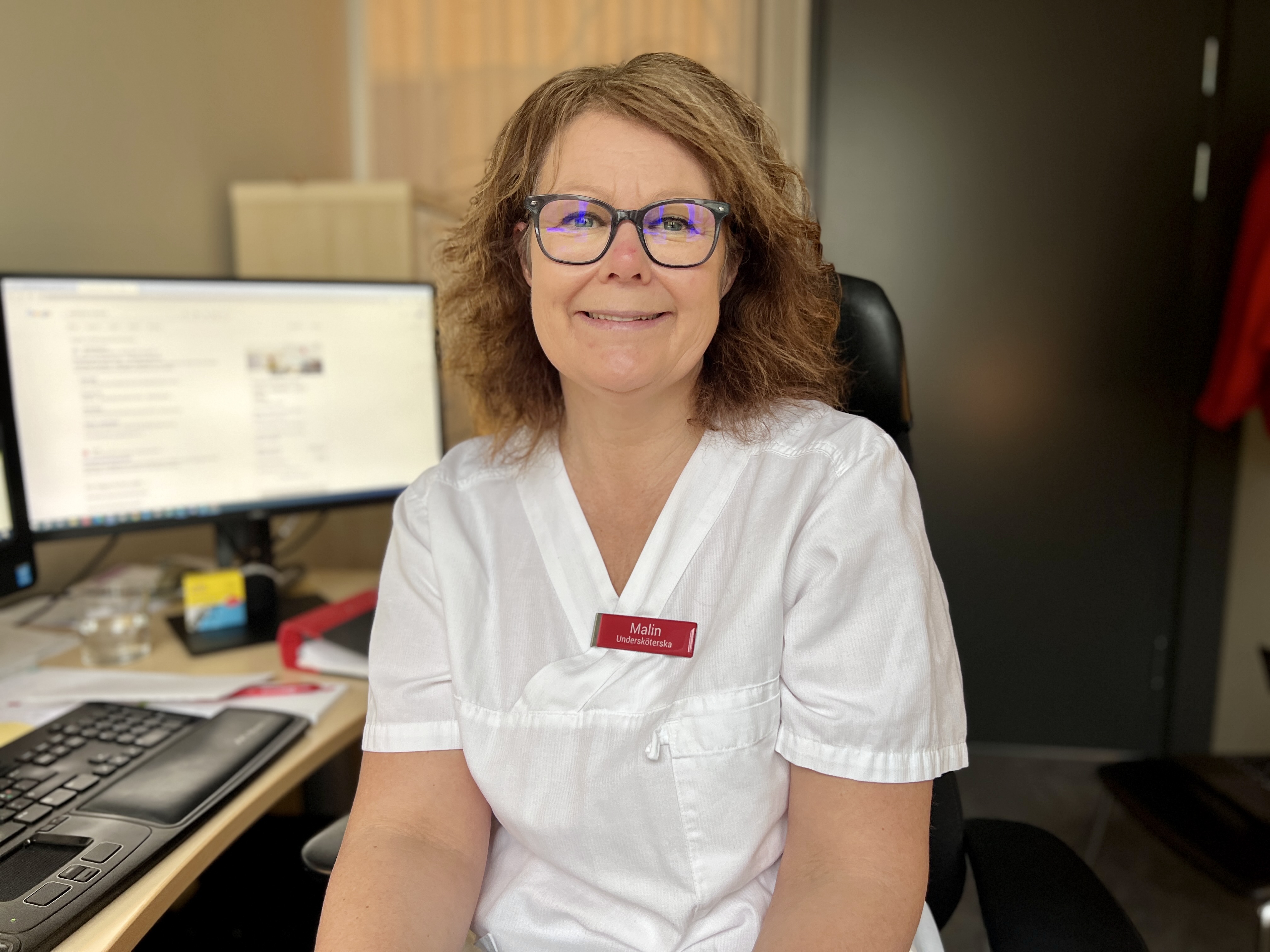 En person med glasögon och sjukvårdskläder tittar in i kameran och ler. Det står Malin, undersköterska på hennes namnskylt. 