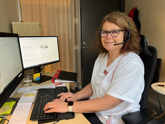 En person i glasögon sitter med headset vid ett tangentbord. Hon tittar mot kameran och ler.  