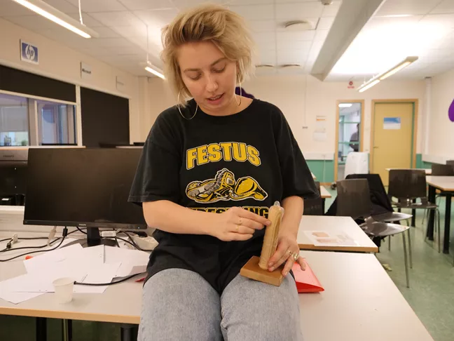 Linda trär en kondom över en träkuk. 
