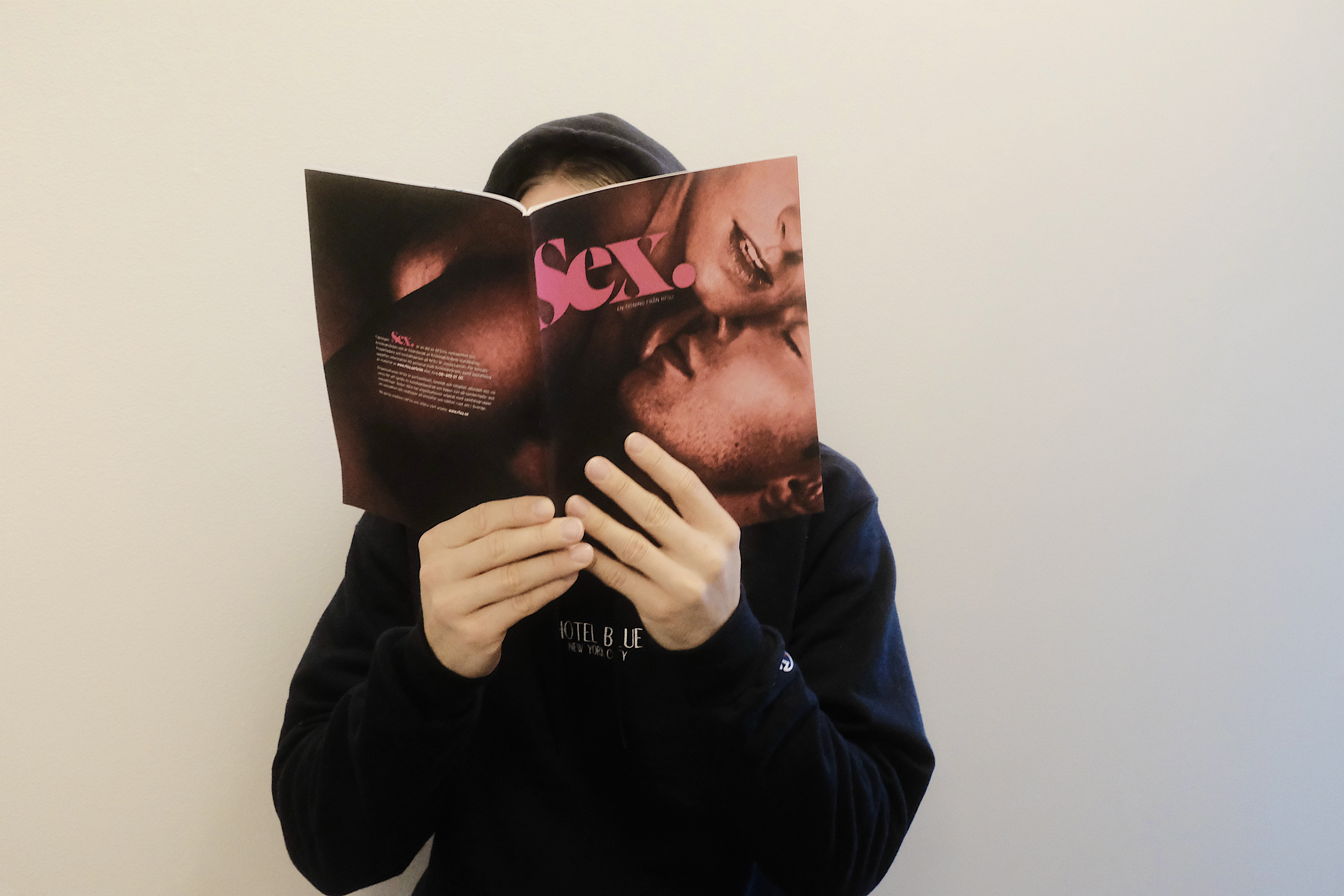 Man sitter mot en vit vägg och läser tidningen “Sex”. Han har en hoodie på sig och ansiktet täcks av tidningen.  