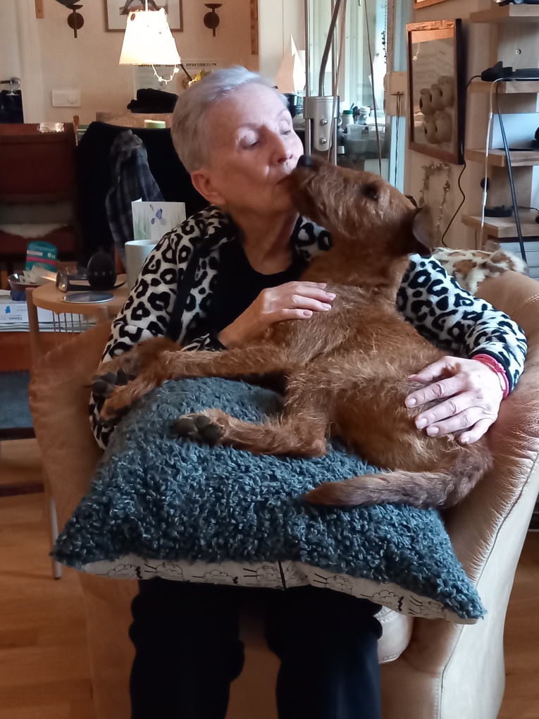 En kvinna i grått hår sitter med sin hund i knäet. De pussar på varandra. 